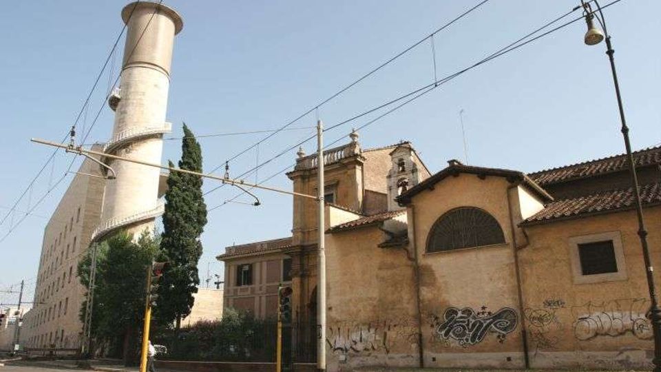 Vista stradale urbana con un grande edificio industriale in muratura e un’alta ciminiera cilindrica bianca, affiancati da un fabbricato basso con tetto in tegole e pareti con graffiti. In primo piano si vedono binari del tram, cavi aerei, pali e un lampione lungo la strada.