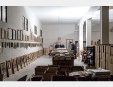 Ampio locale interno tipo deposito o archivio con pareti bianche e soffitto alto, con molti quadri e cornici appoggiati a terra e appesi lungo la parete sinistra. Al centro e in primo piano si vedono tavoli e scatole con pile di documenti e libri, mentre a destra ci sono colonne ad arco e alcuni pianoforti vicino a scaffalature.