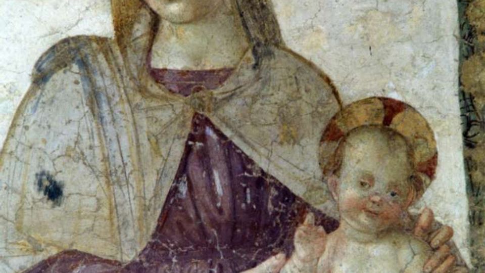 Affresco religioso su parete interna, con figure aureolate dipinte in stile medievale entro una cornice decorativa. Intonaco e pigmenti appaiono consumati e screpolati, evidenziando l’antichità della superficie muraria.