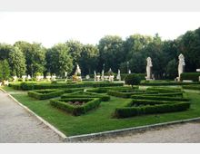 Giardino formale con siepi basse potate a disegni geometrici e vialetti in ghiaia, con ampi prati. Sullo sfondo si vedono statue e vasi ornamentali in pietra davanti a una fitta fila di alberi.