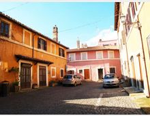Piccola piazza acciottolata in un borgo, circondata da edifici bassi dai colori caldi (arancione e rosa) con persiane, porte in legno e tetti in tegole. Sono presenti lampioni a parete, alcuni vasi di piante e auto parcheggiate lungo i lati.