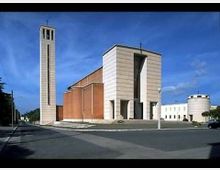 Chiesa moderna con facciata in pietra chiara e navata laterale in mattoni, affiancata da un alto campanile rettangolare con croce in cima. Davanti si apre un’ampia piazza asfaltata con strada e lampioni, con un edificio bianco sullo sfondo sotto un cielo azzurro.
