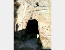 Ingresso a un tunnel scavato nella roccia, con apertura ad arco in una parete di pietra naturale. Sopra l’entrata sono visibili cavi e un faro, con una piccola strada in cemento che conduce all’interno buio.