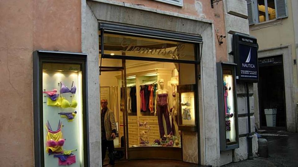 Facciata di un negozio su Via Frattina, con ampia vetrina d’ingresso incorniciata in pietra e due vetrine laterali illuminate con esposizione di capi di lingerie. Intorno si vedono muri intonacati, targhe stradali e insegne, e una strada stretta in contesto urbano storico.