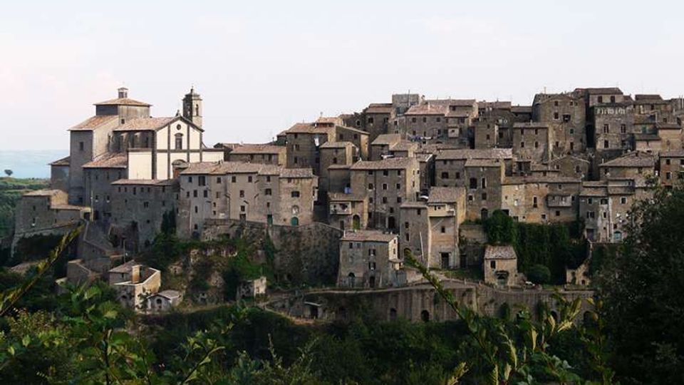 Borgo medievale su un pendio con fitte case in pietra dai tetti in coppi e una chiesa con campanile sul lato sinistro. In primo piano si vedono vegetazione e alberi, con colline sullo sfondo sotto un cielo chiaro.