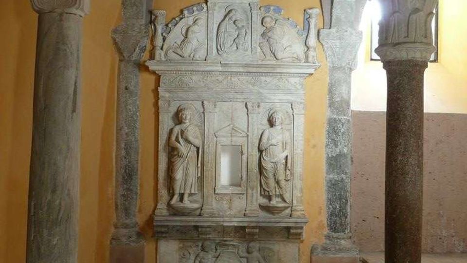 Interno di una chiesa o cappella con volte a crociera e colonne in pietra, pavimento in pietra e pareti color ocra. Al centro si vede un altare o monumento marmoreo riccamente scolpito con rilievi e figure, incorniciato da archi e pilastri.