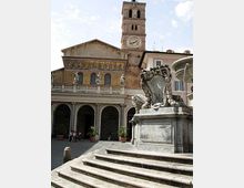 Piazza con scalinata in primo piano e fontana monumentale in pietra decorata. Sullo sfondo si vede una chiesa con portico ad archi, facciata con mosaico e statue, e un campanile con orologio.