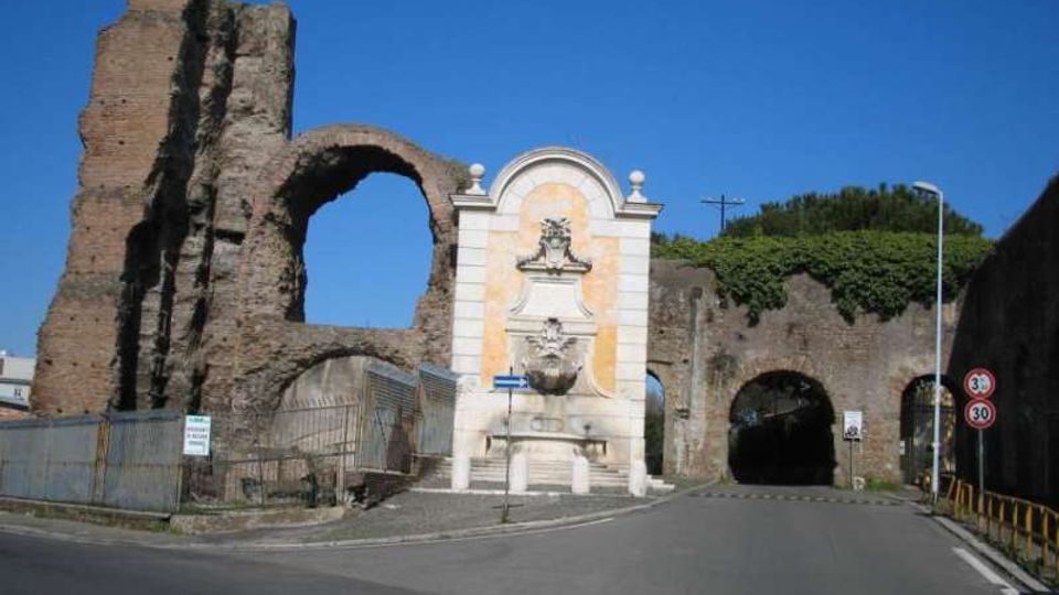 Fontana monumentale addossata a un’antica struttura in mattoni con grandi archi e tratti in rovina, lungo una strada asfaltata. Sullo sfondo si vedono un passaggio ad arco che attraversa il muro e vegetazione in cima alle mura sotto un cielo sereno.