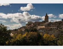 Veduta di un borgo collinare con case in pietra e tetti in coppi, dominato da un campanile con cuspide. In primo piano si vede una fascia di alberi e vegetazione, con cielo azzurro e nuvole sullo sfondo.