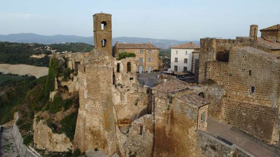 Veduta panoramica di un borgo medievale in pietra con resti di mura e una torre campanaria, affacciato su uno sperone roccioso. Sullo sfondo si vedono colline e campagna, con case compatte attorno a una piccola piazza.
