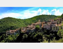 Veduta panoramica di un borgo collinare con case in pietra dai tetti in coppi, raggruppate su un pendio e circondate da fitti boschi. Sullo sfondo si vedono colline verdi e un cielo azzurro con nuvole.
