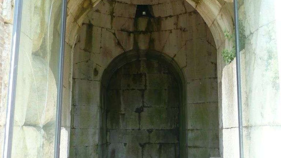 Interno di un passaggio in pietra con volta a botte e pareti di grandi blocchi, che conduce a una nicchia ad arco sul fondo. Ai lati si vedono pannelli di vetro incorniciati e sul pavimento lastre di pietra con piccole chiazze di vegetazione.