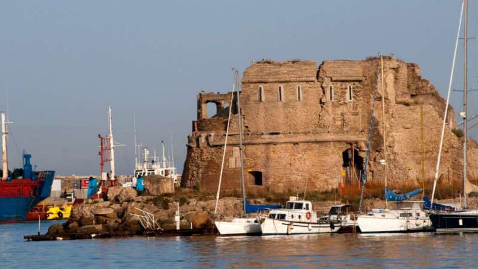 Rovina di una grande costruzione in pietra su un molo, con pareti parzialmente crollate e piccole aperture, affacciata su un porto. In primo piano si vede l’acqua con barche a vela e piccole imbarcazioni ormeggiate, mentre sullo sfondo sono presenti altre navi e strutture portuali.