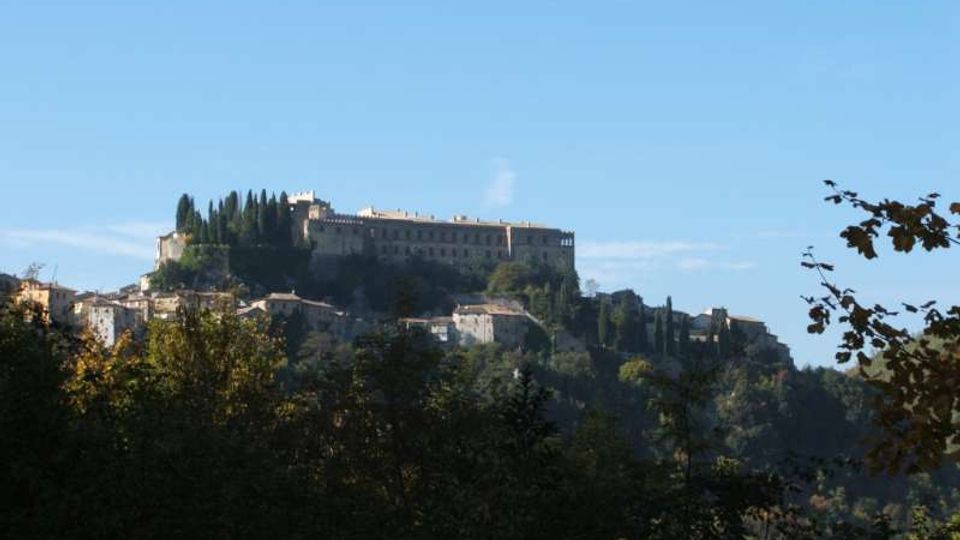 Panorama di un borgo su una collina, con un grande edificio storico fortificato in cima e file di cipressi lungo il crinale. In primo piano si vede un’area boschiva, con case in pietra sparse sui pendii sotto il castello.