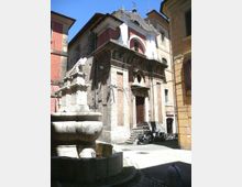 Piccola piazza urbana con una fontana in pietra in primo piano e una chiesa storica sullo sfondo, con facciata in pietra e mattoni, portale centrale e frontone curvilineo. Intorno si vedono edifici alti e ravvicinati lungo una strada stretta, con alcune moto parcheggiate vicino all’ingresso.