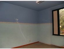 Interno di una stanza vuota con pareti bicolori azzurre, fascia decorativa orizzontale e pavimento grezzo. A destra c’è una finestra con inferriata e telaio scuro, con vista su alberi all’esterno.