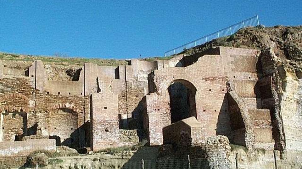Rovine di un complesso in mattoni incassato in una parete di tufo, con grandi arcate e nicchie scavate nella roccia. In alto si vede il bordo del pendio con una ringhiera, sotto un cielo azzurro.