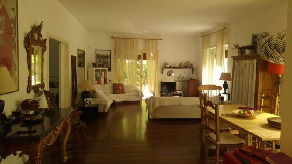 Ampio soggiorno con pavimento in legno scuro, divani chiari e zona pranzo con tavolo e sedie in legno. Grandi finestre e una portafinestra con tende leggere illuminano la stanza, arredata con mobili classici, TV su mobile basso e specchi e quadri alle pareti.