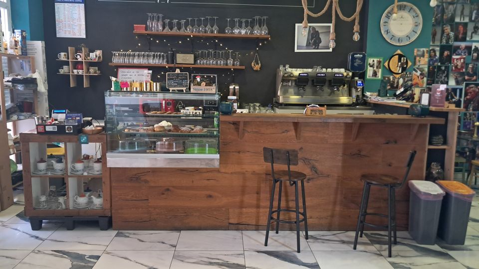 Interno di un lounge bar/caffè con lungo bancone in legno, vetrina per dolci, macchina da espresso e mensole con bicchieri su una parete nera con scritta “Apperò”. Pavimento in piastrelle effetto marmo, sgabelli alti e parete laterale decorata con foto e un grande orologio.
