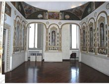 Sala storica con pavimento in legno scuro, pareti decorate da nicchie ad arco con figure dipinte e cornici dorate, e grandi finestre che illuminano l’ambiente. In alto, una fascia di affreschi corre sotto la volta, mentre l’arredo è minimo con una sedia e alcuni piccoli supporti.