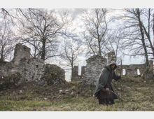 Rovine di un edificio in pietra con muri parzialmente crollati su un pendio erboso, circondato da alberi spogli. Sullo sfondo si vede un cielo nuvoloso sopra i resti delle strutture.