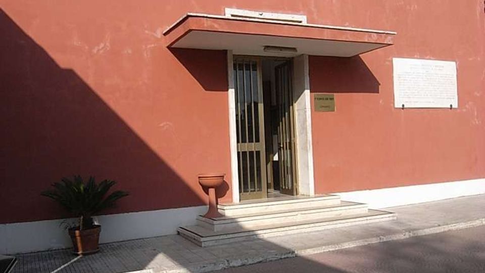 Ingresso di un edificio basso con facciata intonacata color rosso mattone, piccola pensilina e gradini in marmo che conducono a una porta vetrata con inferriata. Davanti ci sono un vialetto pavimentato, una piccola aiuola con blocco in pietra e una targa informativa sulla parete.