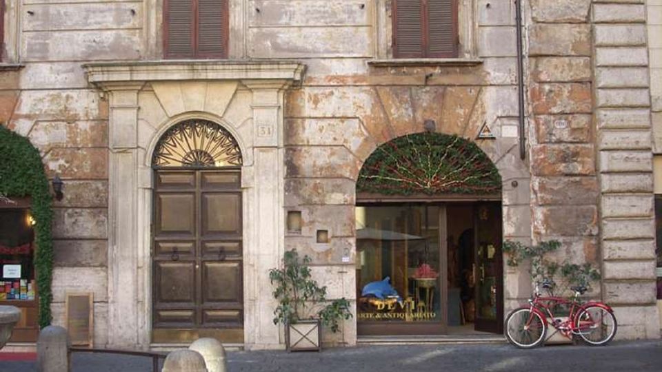 Facciata di un edificio storico in pietra intonacata, con grandi portoni e finestre con persiane scure, un piccolo balcone in ferro e due ingressi ad arco al piano terra. Davanti si vede una strada lastricata con dissuasori in pietra, alcune piante in vaso e una bicicletta appoggiata vicino all’ingresso.