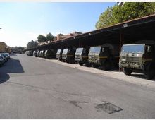 Ampio piazzale asfaltato in un deposito veicoli, con una lunga fila di camion militari parcheggiati sotto una tettoia metallica con copertura rossa. Sullo sfondo si vedono edifici e alberi lungo il perimetro dell’area.