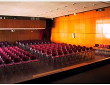 Sala auditorium con file di sedie bordeaux disposte su pavimento in legno scuro, con pareti rivestite in pannelli di legno e illuminazione a soffitto. Sul lato destro si vede un’area rialzata tipo palco e, in fondo, una tenda scura vicino all’ingresso.