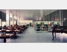 Ampia sala di biblioteca con lunghi tavoli e sedie in legno disposti in file, affiancati da scaffalature metalliche piene di libri. Ambiente moderno con soffitto alto a pannelli, pavimento chiaro e grandi finestre laterali che illuminano lo spazio.