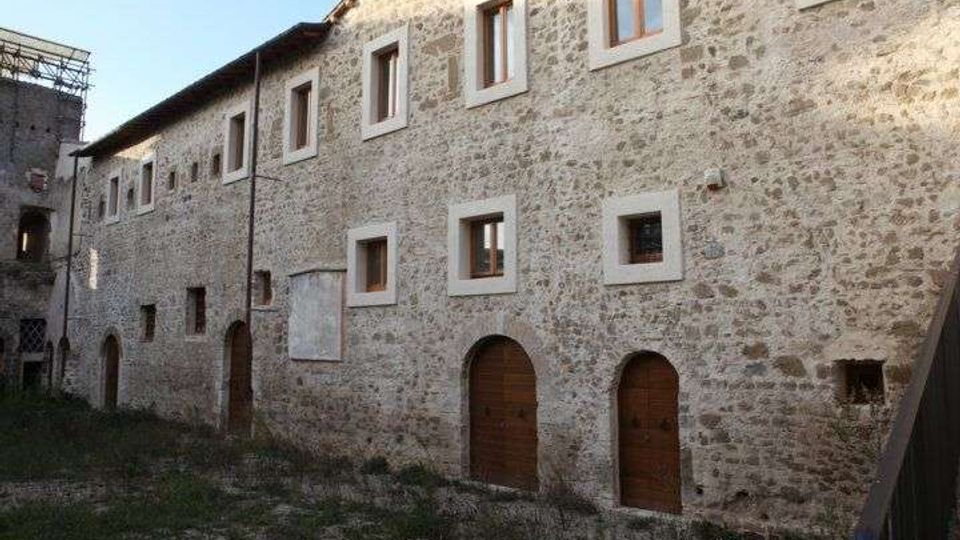 Cortile interno con un lungo edificio storico in pietra a due piani, con finestre rettangolari incorniciate e portali ad arco in legno al piano terra. Il terreno è in parte erboso e l’area è chiusa da altre strutture in muratura sul lato sinistro.