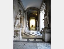 Corridoio monumentale in pietra con archi e colonne, pavimento a scacchi bianco e nero e statue classiche su piedistalli lungo entrambi i lati. In fondo si apre un vano luminoso con una statua centrale, illuminato da luce naturale.