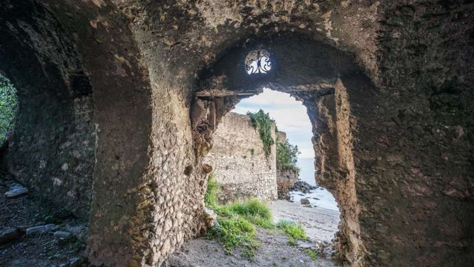 Interno in pietra di una struttura antica con volta ad arco, che inquadra un’apertura irregolare verso l’esterno. Attraverso il varco si vedono mura in rovina, vegetazione e una spiaggia rocciosa sul mare.