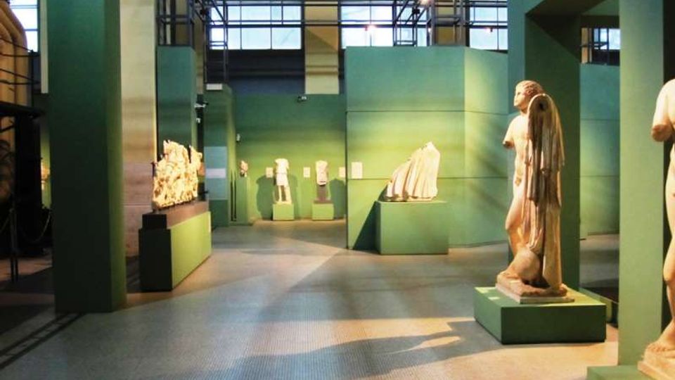 Interno di un museo o spazio espositivo con pareti e piedistalli verde acqua che ospitano statue e frammenti scultorei. L’ambiente è ampio e industriale, con soffitto alto in metallo e grandi finestre a griglia che illuminano la sala.
