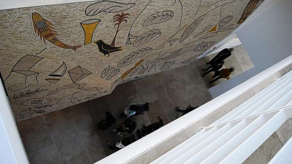 Vista dall’alto di un atrio interno di edificio moderno con pavimento in pietra e pareti bianche, attraversato da una passerella. Su una grande parete laterale è presente un ampio murale astratto con simboli e forme stilizzate in colori ocra e nero.