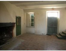 Interno di una stanza spoglia con pareti intonacate chiare e pavimento in mattoni, con grande camino in muratura su un lato. Sono visibili una finestra e due porte in legno verde, una delle quali rialzata su alcuni gradini.