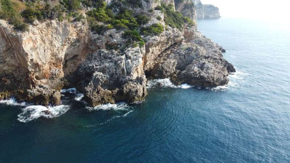 Scogliera costiera rocciosa con pareti di pietra e vegetazione mediterranea, affacciata su mare blu. Onde che si infrangono sugli scogli e promontorio che si estende verso l’acqua.