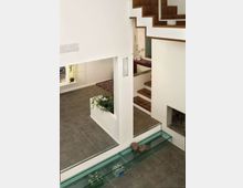 Interno di una casa moderna su più livelli con pareti bianche, pavimento in grandi piastrelle grigie e scale in legno con parapetto bianco. In primo piano c’è una lunga canaletta a filo pavimento in vetro trasparente, con elementi decorativi e piante.