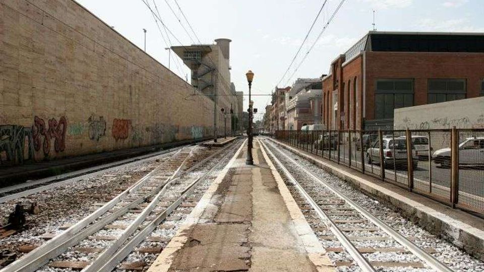 Binari del tram in un corridoio urbano con banchina centrale stretta e lampione, sotto cavi aerei. Ai lati si vedono muri con graffiti, edifici industriali e una recinzione con auto parcheggiate.