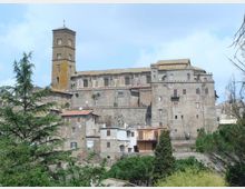 Complesso storico in pietra su un’altura, con un alto campanile quadrato e edifici fortificati con piccole finestre e tetti in coppi. In primo piano si vedono case in muratura e alberi, con vegetazione e cielo sullo sfondo.