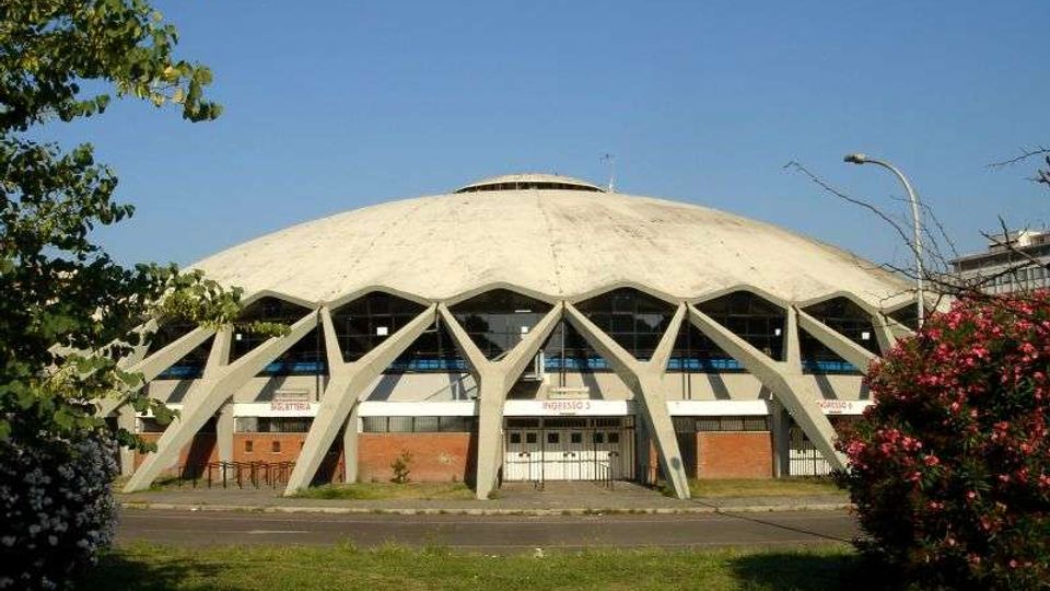 Edificio moderno a cupola, simile a un palazzetto dello sport, con grandi archi in cemento a vista lungo la facciata e ingresso centrale con vetrate. Davanti si vede una strada con prato e alberi e cespugli ai lati, sotto un cielo sereno.