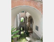 Passaggio stretto in un borgo, con scala in pietra e pavimento con muschio, coperto da un grande arco e affiancato da portici ad arcate. Sulle pareti intonacate si vedono una piccola balconata con ringhiera in ferro battuto e aperture ad arco che conducono a cortili interni.