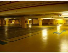Parcheggio sotterraneo vuoto con ampie corsie asfaltate e segnaletica a terra, inclusa una grande freccia direzionale blu. Colonne in cemento numerate e illuminazione al neon sul soffitto basso con tubazioni a vista.