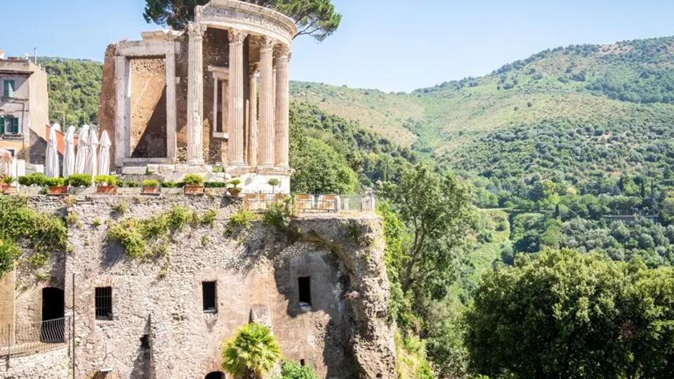 Rovine di un tempio circolare in pietra con colonne classiche su un promontorio roccioso, con un grande pino che sovrasta la struttura. Sullo sfondo si vedono colline verdi e una valle boscosa sotto un cielo sereno.