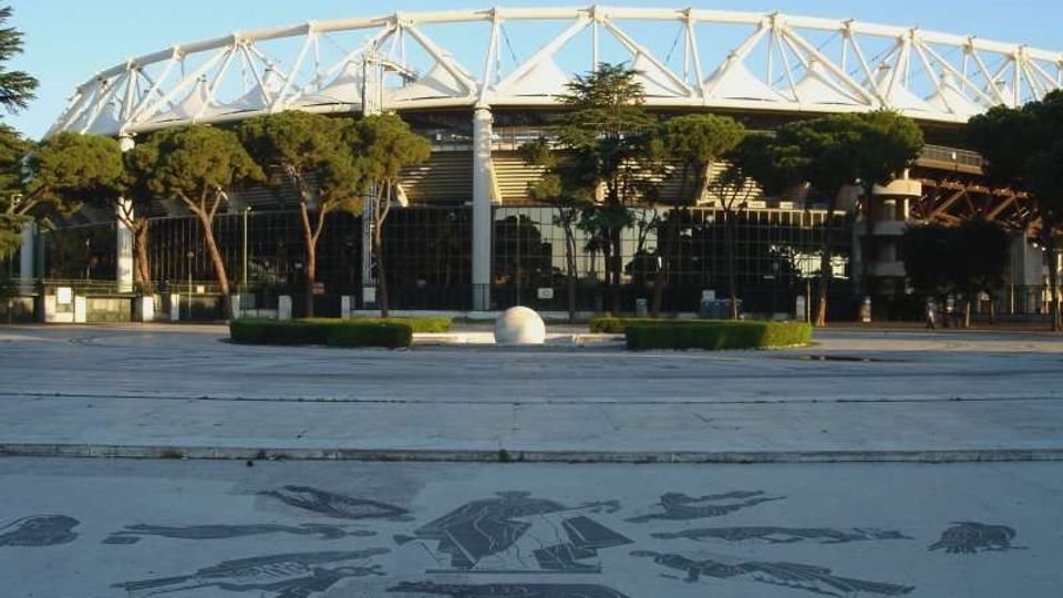 Veduta esterna di un grande stadio moderno con struttura ad anello e travi bianche a traliccio sul tetto, circondato da alberi e facciate vetrate. In primo piano si vede un’ampia piazza pavimentata con decorazioni a mosaico e aiuole basse.