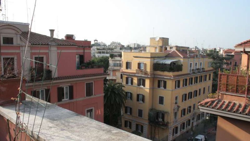 Veduta urbana da una terrazza con muretto in primo piano, affacciata su edifici residenziali di più piani con facciate colorate e tetti in coppi. Sullo sfondo si vede lo skyline della città con altri palazzi e alberi lungo la strada sottostante.
