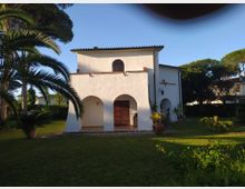 Edificio bianco in stile mediterraneo con portico ad archi e tetto in tegole, affacciato su un prato con palme e piante in vaso. Sul lato destro si vede una croce sulla facciata e un piccolo ingresso ad arco, circondati da alberi alti.