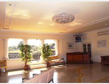 Ampia sala interna in stile classico con grandi finestre ad arco e ringhiera decorativa, pavimento a mosaico e lampadario centrale. Sono presenti panche e divani lungo le pareti, piante in vaso vicino alle finestre e un bancone in legno in un angolo.