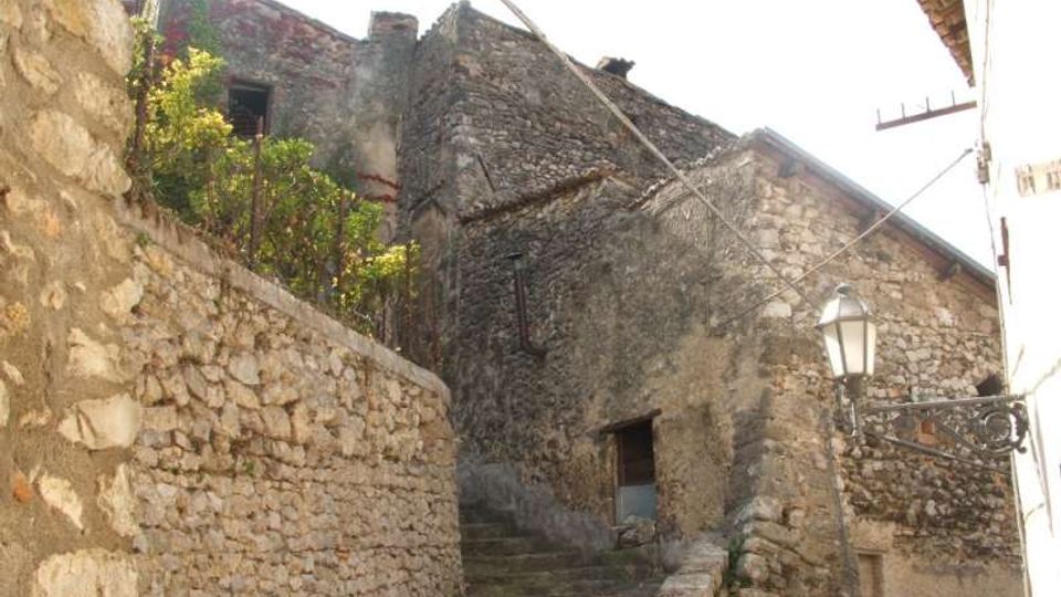 Scalinata in pietra in un vicolo stretto di un borgo antico, affiancata da alti muri e case in muratura grezza. Si vedono una ringhiera in ferro battuto, un lampione a parete e cavi aerei tra gli edifici.