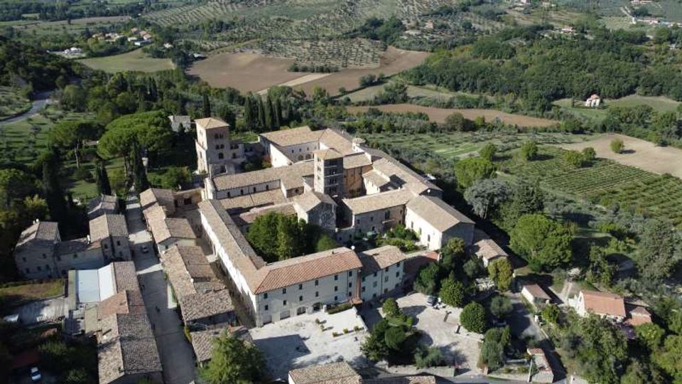Veduta aerea di un grande complesso monastico in pietra con tetti in coppi, disposto attorno a cortili interni e una torre centrale. L’edificio è circondato da alberi, giardini e campi coltivati in una campagna collinare.